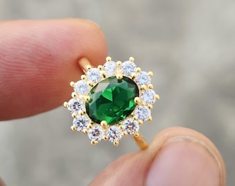 May include: A gold ring featuring a large, oval-cut, deep green center stone surrounded by a halo of twelve small, round, clear white stones. The setting is crafted from a polished yellow metal, creating a bright contrast with the green gem.