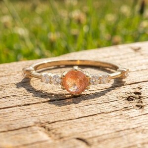 Puede incluir: Un anillo de oro con una piedra de sol redonda de color melocotón en el centro, flanqueada por dos pequeñas piedras redondas transparentes a cada lado. La banda es fina y metálica, con un acabado pulido que refleja la luz.