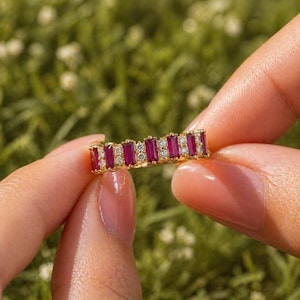 May include: A gold ring featuring alternating rectangular rubies and small round diamonds. The ring is held between two fingers against a blurred green background, showcasing the jewelry's vibrant red and sparkling white stones.