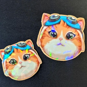 以下が含まれることがあります： 2匹の猫の形をしたステッカー。猫はオレンジと白の毛並みで、緑色の目をし、青いアビエーターゴーグルをかけています。ステッカーはホログラフィックで虹色の仕上がりで、金の縁取りがあります。背景は黒です。