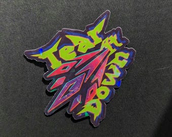 May include: A holographic sticker with the words "Tear It Down" in neon green and pink. The sticker has a jagged, abstract design with a dark background.