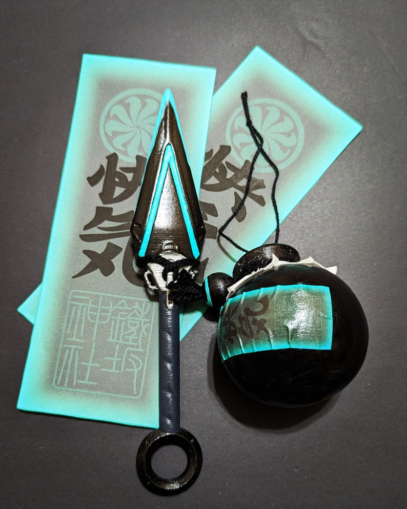 Peut inclure: Une arme de ninja noire et bleue avec une poign&eacute;e en anneau et un r&eacute;cipient rond bleu et noir avec des caract&egrave;res japonais. L'arme et le r&eacute;cipient sont sur une surface grise avec deux &eacute;tiquettes en papier turquoise avec des caract&egrave;res japonais.