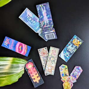 May include: A collection of holographic stickers featuring various designs, including a donut, a ticket, a star, and a flame. The stickers are iridescent and have a rainbow effect.