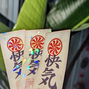 May include: Three iridescent stickers with a red and white circular design and Japanese characters. The stickers have a holographic finish and are arranged in a stack.