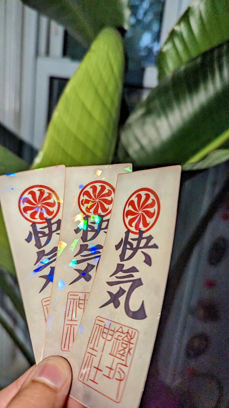 May include: Three iridescent stickers with a red and white circular design and Japanese characters. The stickers have a holographic finish and are arranged in a stack.