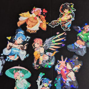 Op de afbeelding: Een verzameling van negen holografische stickers met diverse anime-stijl personages in dynamische houdingen. De ontwerpen bevatten figuren met gekleurd haar, vleugels, paraplu's en muzieknoten, allemaal uitgevoerd met een glinsterende, prismatische afwerking.