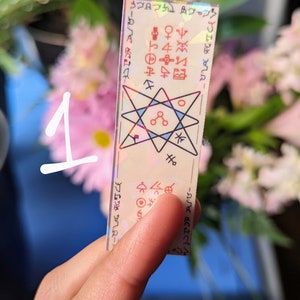 May include: A holographic sticker with a six-pointed star design and various symbols. The sticker is on a white background and has a rainbow iridescent finish.