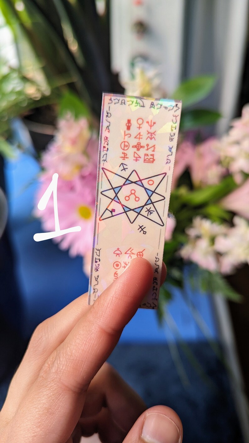 May include: A holographic sticker with a six-pointed star design and various symbols. The sticker is on a white background and has a rainbow iridescent finish.