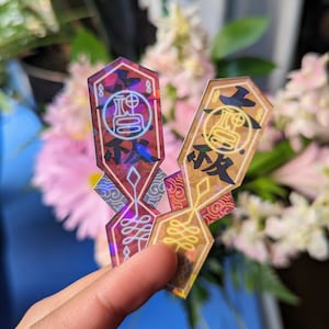 May include: Two iridescent holographic stickers with a geometric design and Japanese characters. The stickers are in shades of red, gold, and blue.