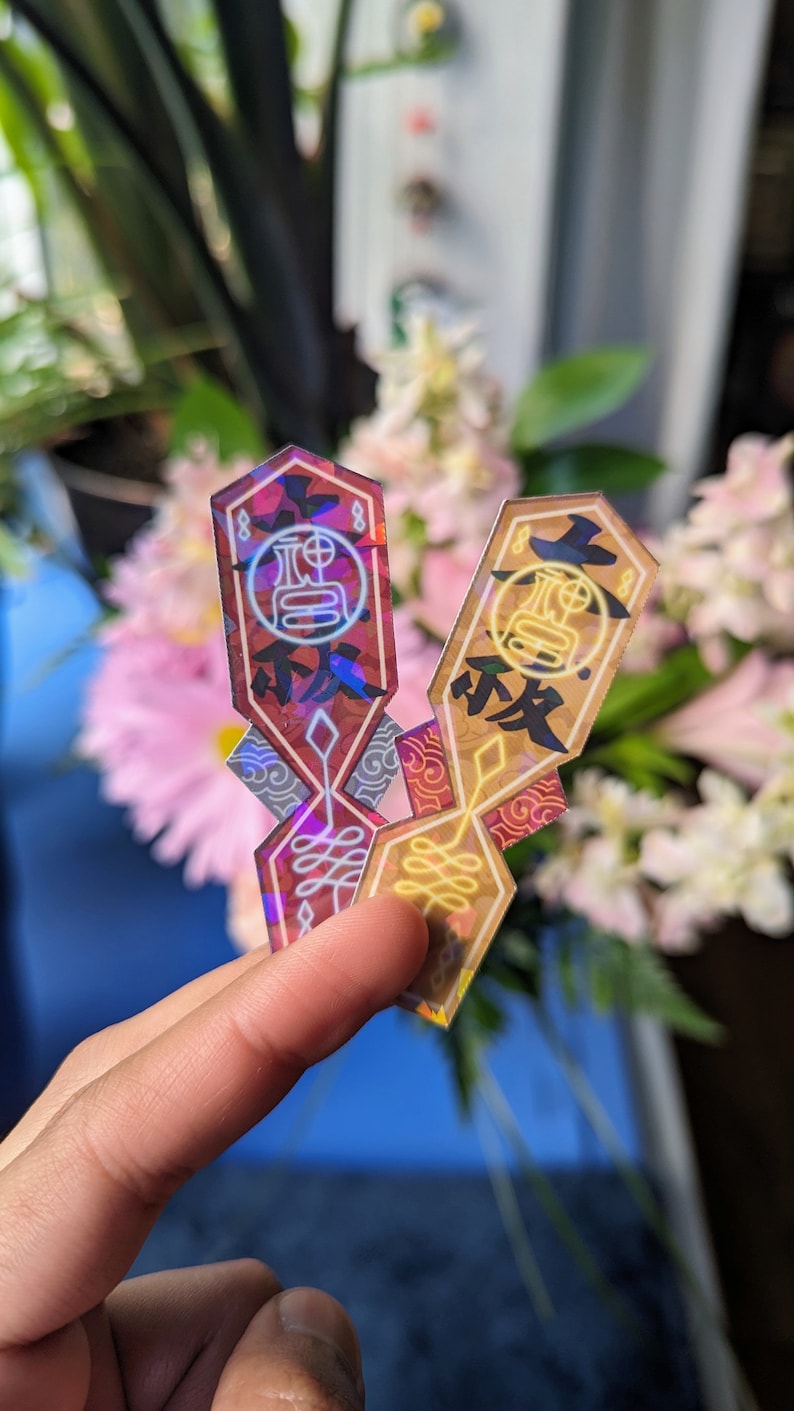 May include: Two iridescent holographic stickers with a geometric design and Japanese characters. The stickers are in shades of red, gold, and blue.