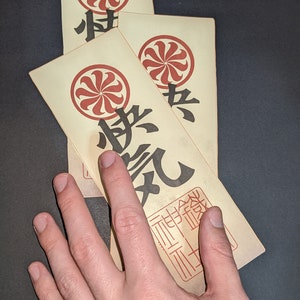 Peut inclure: Trois cartes en papier vintage avec un motif circulaire rouge et blanc et des caract&egrave;res japonais noirs. Les cartes sont empil&eacute;es les unes sur les autres et la carte du dessus est partiellement cach&eacute;e par une main.