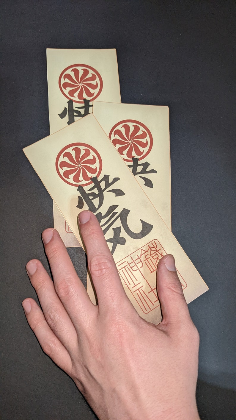 Peut inclure: Trois cartes en papier vintage avec un motif circulaire rouge et blanc et des caract&egrave;res japonais noirs. Les cartes sont empil&eacute;es les unes sur les autres et la carte du dessus est partiellement cach&eacute;e par une main.