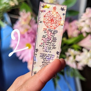 May include: A white card with a red and black symbol in the center. The card has a holographic finish and is surrounded by a black border. The card has text written in a foreign language.