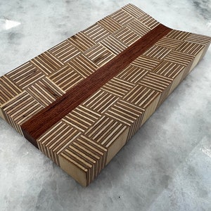 May include: A wooden cutting board with a geometric pattern made from different types of wood. The board has a reddish-brown stripe running down the center.