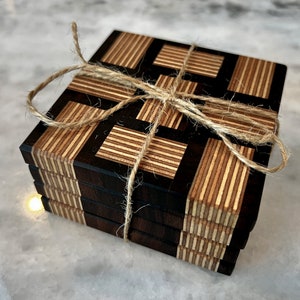 May include: A set of four coasters made of wood and black resin. The coasters are stacked and tied together with twine. The coasters have a geometric pattern of black and wood stripes.