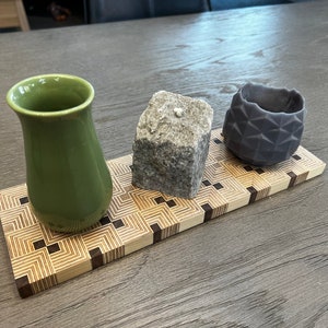 May include: A wooden tray with a geometric pattern features a green ceramic vase, a gray stone, and a dark gray ceramic bowl.