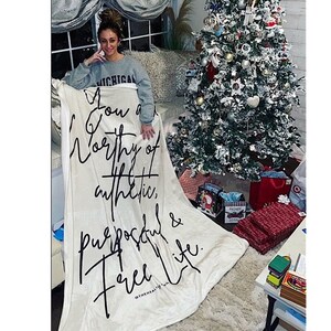 Puede incluir: Una manta blanca de felpa con texto negro que dice "You are worthy of authentic, purposeful & free life." La manta está colocada sobre un sofá con una persona sentada en él. Hay un árbol de Navidad en el fondo.