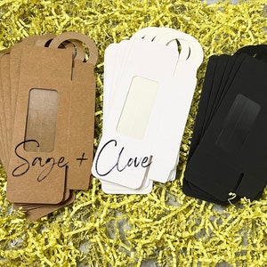 May include: A collection of gift tags in three colors: kraft brown, white, and black. Each tag has a rectangular window and a circular hole for hanging. The kraft brown tags read "Sage + Clove" in script.