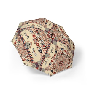 以下が含まれることがあります： 赤、青、白の色の幾何学模様が施されたベージュの傘。模様は伝統的な刺繍を思わせる。