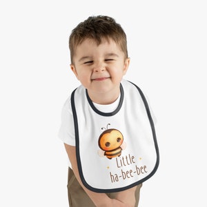 Könnte beinhalten: Ein weißer Baby-Lätzchen mit schwarzem Rand zeigt eine Cartoon-Biene mit dem Text "Little ha-bee-bee".
