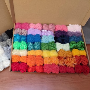 Puede incluir: Una caja llena de ovillos de lana de colores en varias texturas, incluyendo peludas, brillantes y lisas. La lana está dispuesta en filas y está lista para proyectos de manualidades.