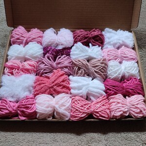 May include: A collection of 20 skeins of yarn in various shades of pink, white, and red. The yarn is arranged in a box, with each skein neatly placed side by side.