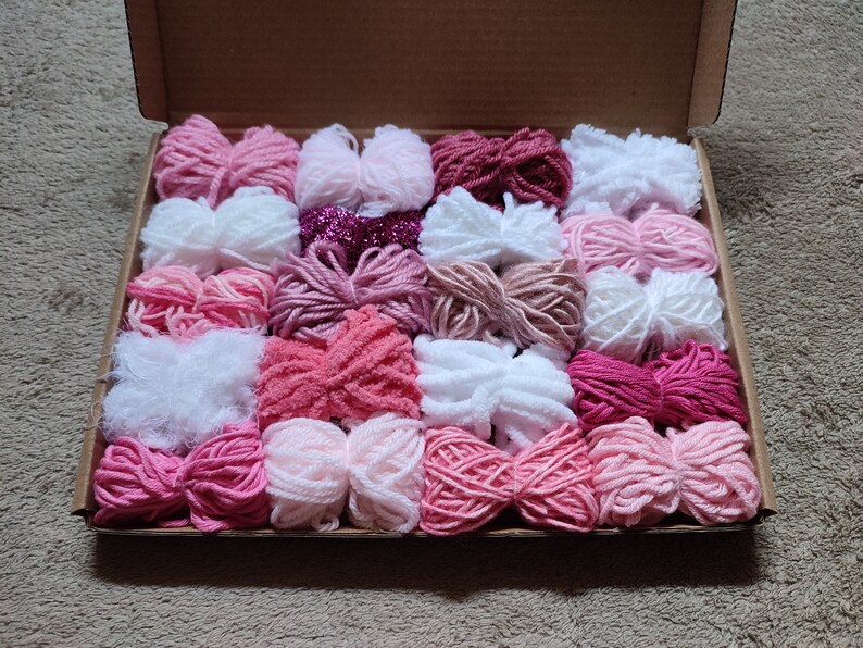 May include: A collection of 20 skeins of yarn in various shades of pink, white, and red. The yarn is arranged in a box, with each skein neatly placed side by side.
