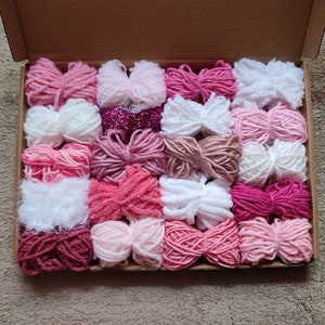 May include: A box filled with 24 skeins of yarn in various shades of pink, white, and red. The yarn is arranged in rows and is ready for crafting projects.