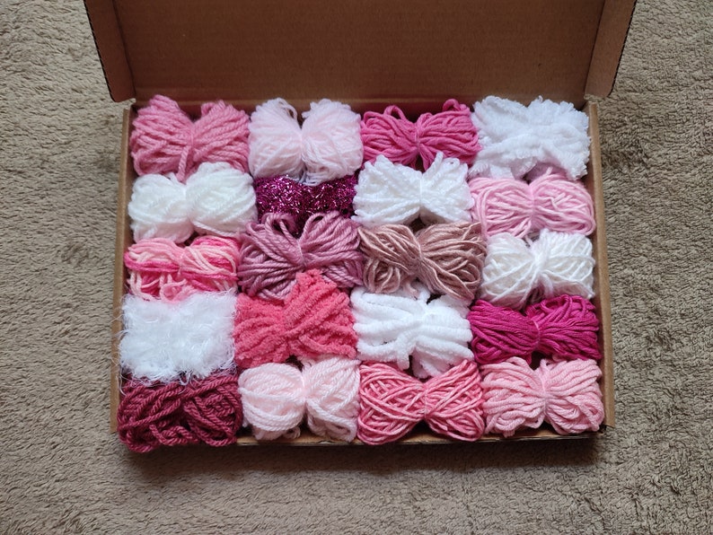 May include: A box filled with 24 skeins of yarn in various shades of pink, white, and red. The yarn is arranged in rows and is ready for crafting projects.