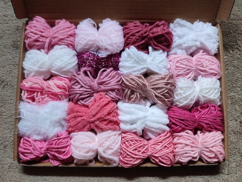 May include: A box filled with 20 balls of yarn in various shades of pink, white, and red. The yarn is arranged in rows and is ready for crafting projects.