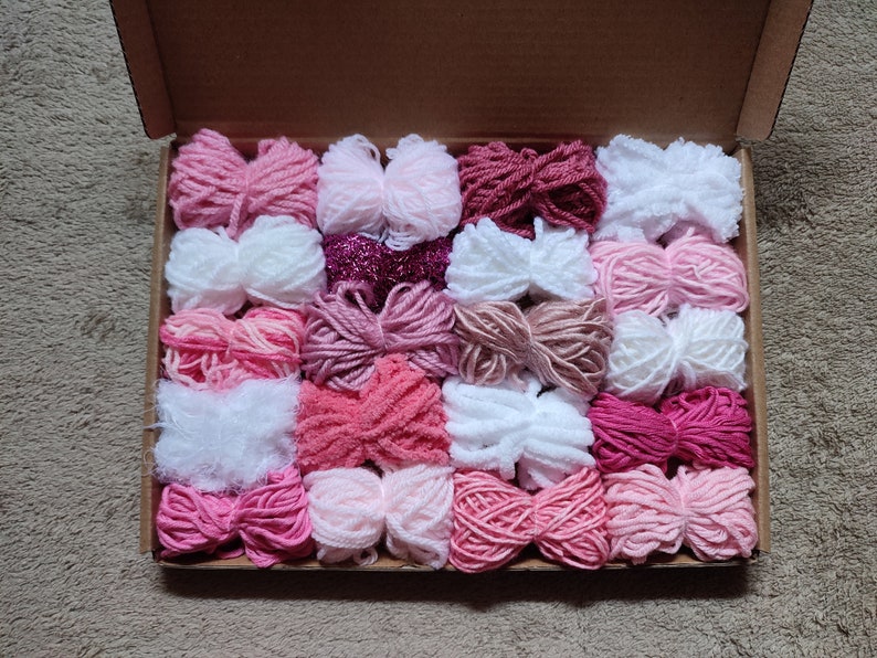 May include: A box filled with various shades of pink, white, and red yarn. The yarn is arranged in rows and is ready for crafting projects.