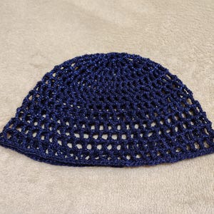 May include: A navy blue crocheted hat with an open weave design. The hat is made of a shiny, textured yarn and is shown on a neutral background. The hat has a rounded crown and a slightly flared brim.