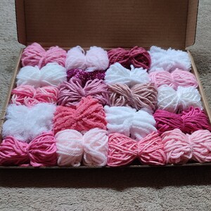 May include: A box filled with pink, white, and red yarn balls. The yarn is arranged in rows and is ready to be used for knitting or crocheting.