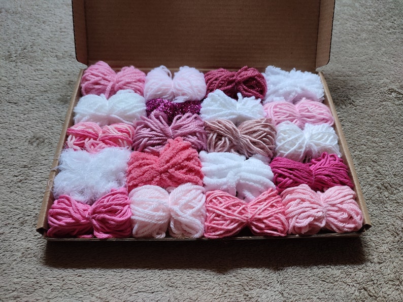May include: A box filled with pink, white, and red yarn balls. The yarn is arranged in rows and is ready to be used for knitting or crocheting.