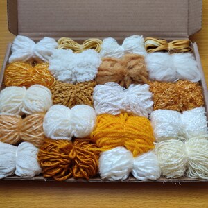 Puede incluir: Una caja llena de 20 ovillos de hilo en varios tonos de blanco y amarillo. El hilo está dispuesto en filas y está listo para proyectos de manualidades.