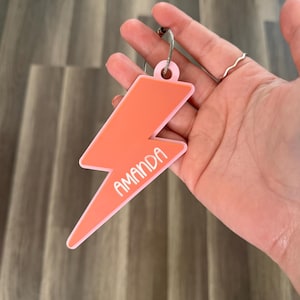May include: A coral-colored lightning bolt-shaped keychain with a light pink border and a silver-colored metal loop. The name "AMANDA" is written in white script across the front of the lightning bolt.