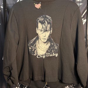 May include: Dark gray long-sleeved shirt featuring a portrait of an actor with the text "Cry-Baby" below the image. A pink heart with an arrow is embroidered on the upper back. The shirt has a high collar and a cropped design.