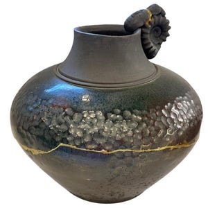 May include: A dark, rounded ceramic vase with a matte black neck and a textured, iridescent body featuring shades of blue, green, and purple. A gold-colored line encircles the lower section, and a spiral-shaped handle sits on the rim.