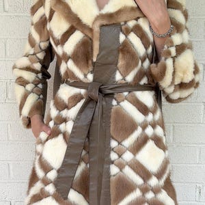 May include: A mid-length coat featuring a geometric diamond pattern in alternating cream and brown fur panels. The garment includes a wide collar, long sleeves, and a solid brown leather-like belt tied at the waist, with visible side pockets.