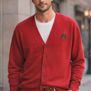 May include: A bright red V-neck cardigan sweater featuring a button-down front and a small embroidered crest logo on the left chest. The garment is worn over a plain white crew neck t-shirt, paired with beige trousers and a brown leather belt.