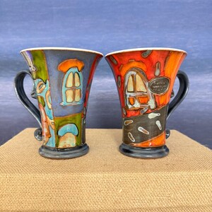May include: Two ceramic mugs with flared rims and dark handles. The left mug features a blue, green, and orange house design. The right mug displays an orange and black house motif with a small oval plaque containing illegible text.