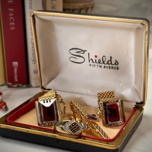 May include: A set of gold-tone jewelry including two rectangular cufflinks with deep red stones and a matching ring with a square textured inlay, all presented in a black and gold-tone hinged box labeled Shields Fifth Avenue.