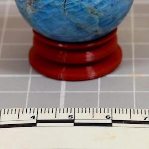 Könnte beinhalten: Eine blaue Edelstein-Kugel mit strukturierter Oberfläche, die auf einem kleinen, roten Holzständer ruht. Die Kugel hat ein marmoriertes Aussehen mit helleren blauen und grauen Adern. Der Ständer ist rund mit einem abgestuften Sockel. Der Hintergrund ist eine Holzoberfläche.