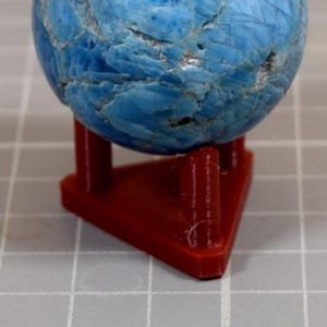 Op de afbeelding: Een blauwe bol, mogelijk een edelsteen, staat op een kleine rode standaard. De bol heeft een textuur met verschillende blauwtinten. De standaard heeft een eenvoudige driehoekige vorm.