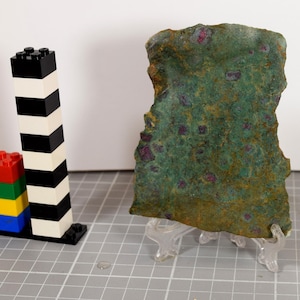 Op de afbeelding: Een groen getinte steen met rode insluitsels, gepresenteerd op een transparante standaard. Daarnaast twee stapels kleurrijke speelgoedbouwstenen, één zwart-wit, de andere rood, groen, geel en blauw. De achtergrond is wit.
