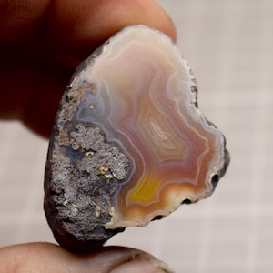 Puede incluir: Una piedra de ágata pulida con forma de corazón. La piedra presenta bandas de color, incluyendo blanco, melocotón, rojo y amarillo. El borde exterior es áspero y gris oscuro. La piedra se sostiene entre los dedos.