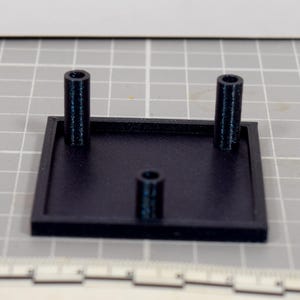 May include: A black, 3D-printed square base with three cylindrical posts. Two posts are on opposite sides, and one is centered. The base is on a gray grid surface with a ruler at the bottom.
