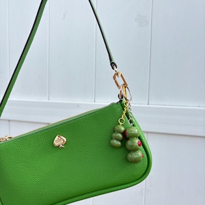 May include: A bright green textured leather shoulder bag with a gold-tone spade logo emblem. The bag features a single strap and a gold-tone zipper closure. Attached to the strap hardware is a decorative keychain featuring two charms shaped like olives.