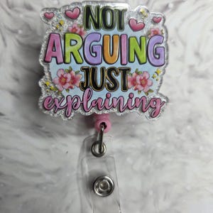 May include: A badge reel with the text "NOT ARGUING JUST explaining" in colorful letters, surrounded by pink hearts and flowers. The badge reel has a clear plastic holder and a pink retractable cord.