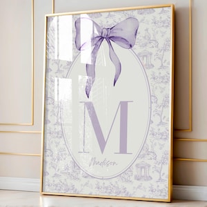 May include: A framed print with a white background and a purple bow at the top. The print features a large purple letter "M" in a circle with the name "Madison" written below it. The background is a repeating pattern of small, light purple trees and houses.